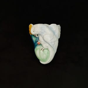 May include: A white ceramic parrot figurine with a yellow crest, perched on a blue and green branch. The parrot has a red accent and a light blue body. The figurine is approximately 13 cm tall.