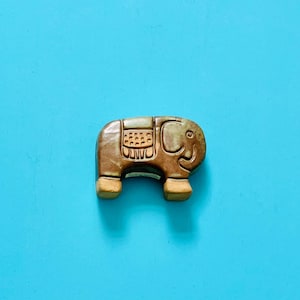 以下が含まれることがあります： 詳細なデザインの小さな茶色の象の置物。象は笑顔で、明るい青色の背景に配置されています。置物の長さは約5cmです。