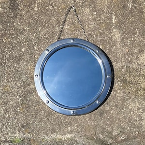 May include: A circular mirror with a silver-toned, riveted frame, hanging from a chain. The mirror reflects the sky. The frame has a nautical or industrial design. The mirror's diameter is approximately 30 cm.