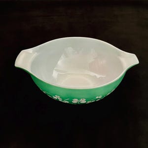May include: A round, green glass bowl with two integrated side handles. The exterior features a repeating white pattern of three-leaf clovers encircling the base. The interior of the bowl is smooth, glossy, and white, contrasting with the vibrant green exterior finish.
