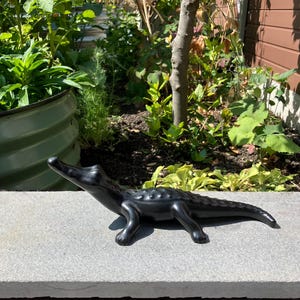 May include: A matte black ceramic figurine of an alligator in a crawling position. The sculpture features a smooth, elongated body with a textured back, a raised snout, and four legs positioned on a flat, light grey stone surface in an outdoor setting.