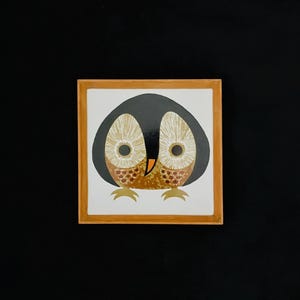 May include: Square ceramic tile with a stylised owl design. The owl has a black head, large beige eyes, and a brown and beige body. The tile is framed in a light brown wooden frame, set against a black background.