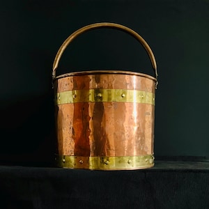 May include: A copper bucket with a brass handle and brass bands. The hammered copper body has a cylindrical shape. The brass bands are riveted to the copper. The handle is arched and attached to the top of the bucket.