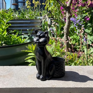 以下が含まれることがあります： グレーの石の表面に置かれた、光沢のある黒い猫型のプランター。猫は微笑んでおり、植物用のバスケットのような質感の容器が付いています。背景には緑豊かな緑とカラフルな花があり、屋外の環境を示唆しています。