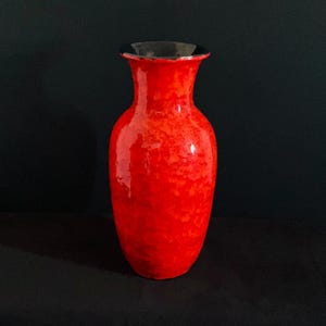 May include: A tall, cylindrical ceramic vase with a vibrant, mottled red glaze featuring varying shades of orange and crimson. The vase has a flared, wide rim with a smooth, dark black interior finish that contrasts with the bright exterior.