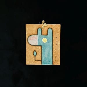 May include: A square ceramic wall hanging with a blue giraffe design on a textured tan background. The giraffe has a pink nose, yellow eye, and a small leaf shape. A beige string is attached for hanging. The piece is likely handmade.