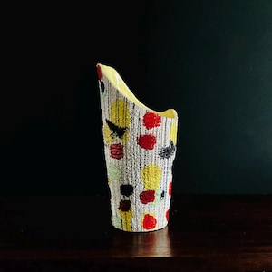 Peut inclure: Un vase en céramique texturé de forme asymétrique. Il est décoré de pois jaunes, rouges, noirs et gris et de lignes verticales sur une base blanche. L'intérieur est jaune pâle.