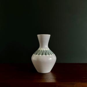 May include: A small, pale grey vase with a flared top and a rounded body. The vase has a decorative band of white and green lines around the middle. The vase is set against a dark green background.