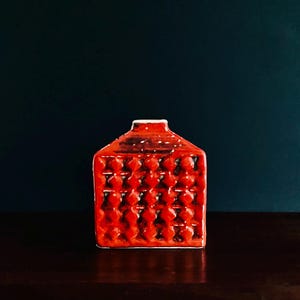 May include: A small, red ceramic vase with a textured, geometric pattern. The vase has a rectangular shape with a narrow neck and a white rim. The vase is displayed against a dark background.