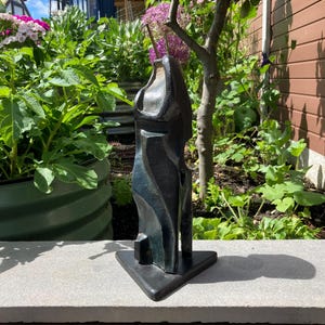 May include: A dark, abstract sculpture with a smooth, reflective surface and curved, organic forms. The piece stands vertically on a flat, triangular base, featuring deep indentations and a polished finish that catches the natural light.