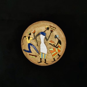 Peut inclure: Une assiette ronde en céramique beige avec une scène peinte de trois personnages stylisés. L'un porte une chemise jaune et un pantalon bleu, un autre une robe blanche et le troisième une tenue rayée. Les personnages semblent tenir des aliments.