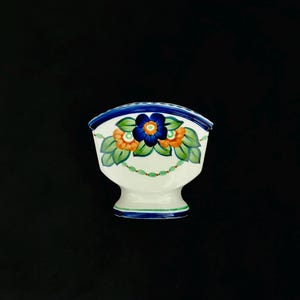 May include: A white ceramic napkin holder featuring a hand-painted floral design with a central dark blue flower, two orange flowers, and green leaves. The piece is accented with a blue rim and a small green beaded garland motif at the base.