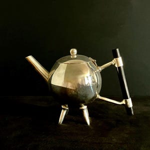 May include: A spherical silver teapot featuring a polished metallic finish, a short angled spout, and a lid with a small round knob. The teapot stands on three short legs and includes a vertical black handle attached by two silver metal arms.