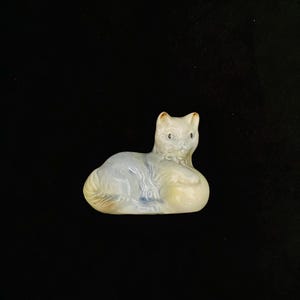 May include: A ceramic cat ornament in a resting pose. The cat is mainly white with light blue and yellow accents. The figurine has a glossy finish and is set against a black background.