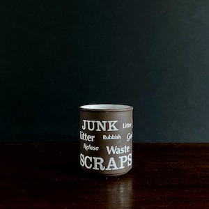 May include: A brown ceramic mug with white text that reads "JUNK, litter, rubbish, refuse, waste, scraps." The mug has a white interior and is set against a dark background. The mug is cylindrical and appears to be handmade.