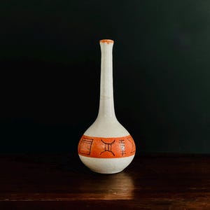 May include: A white ceramic vase with a long neck and rounded base. The vase has an orange band with black geometric designs. The vase is set against a dark background, highlighting its shape and colour.