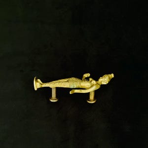 May include: A gold-toned brass cabinet pull shaped like a reclining mermaid figure. The decorative handle features a textured tail, a raised torso, and two cylindrical mounting posts extending from the underside for attachment to furniture.