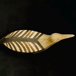 May include: A carved wooden bird decoy featuring a natural light brown finish on the head and beak, with a dark brown and cream-coloured striped feather pattern along the back and wings. The bird has a small, circular red eye detail.