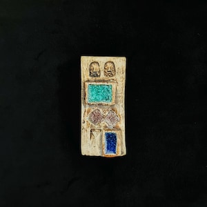 May include: A rectangular ceramic plaque with a textured beige surface featuring two small embossed figures at the top, a bright green rectangular inlay, two diamond-shaped textured inlays, and a deep blue rectangular inlay at the bottom.