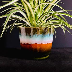 May include: A ceramic plant pot with a glossy finish featuring a layered colour gradient of dark green at the base, transitioning into vibrant orange, white, light blue, and dark green at the rim, containing a spider plant with long green and white leaves.