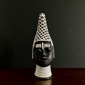Peut inclure: Une sculpture en céramique noire et blanche représentant une tête stylisée. La tête est coiffée d'une coiffe conique et allongée avec un motif en treillis. Le visage est noir avec des détails blancs, et le cou présente des bandes blanches superposées.