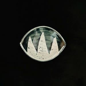 May include: An oval-shaped ceramic dish featuring a dark, mottled blue and grey background. The interior surface is decorated with three white, triangular mountain-like shapes, each detailed with thin, dark, horizontal etched lines.