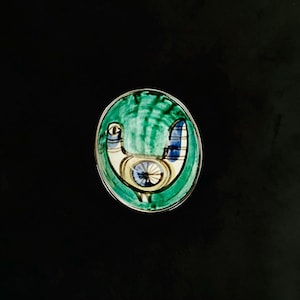 May include: A small, oval-shaped ceramic dish featuring a hand-painted bird motif. The bird is rendered in shades of blue, white, and black, set against a vibrant green background with visible brushstrokes. The dish has a smooth, glossy finish.