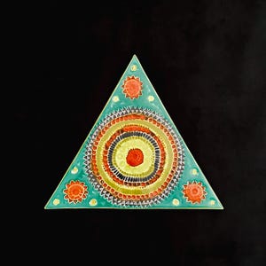 May include: A triangular ceramic tile with a teal border and a central design of concentric circles in red, yellow, and black. The tile features orange sunburst designs at each corner. The background is black.