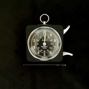 May include: A black Smiths Interval Timer with a circular clock face and a metal ring at the top. The timer has a clear glass cover and white numerals. The timer is approximately 7.6 cm wide.