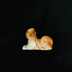May include: A ceramic dog figurine in a reclining position, with brown and white colouring and a glossy finish. The dog is detailed and set against a dark background. A decorative item.