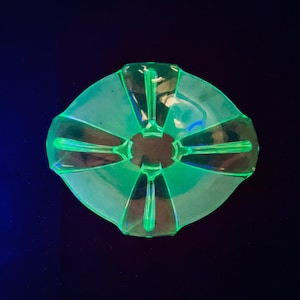 May include: A vibrant, green glass dish with a distinctive starburst pattern. The dish features a central circular area with radiating, elongated segments. The glass has a subtle iridescence, creating a visually appealing contrast against the dark backdrop.
