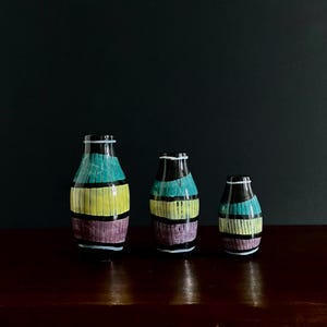 May include: Three decorative vases with a black base and top, featuring horizontal stripes of teal, yellow, and purple. The vases are arranged on a dark wooden surface against a dark background.