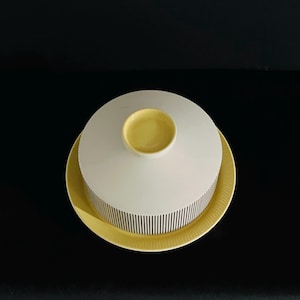 May include: A round butter dish with a white dome lid featuring thin vertical black stripes and a yellow circular knob handle. The base is a matching yellow plate with a ribbed edge, providing a clean and functional design for food storage.