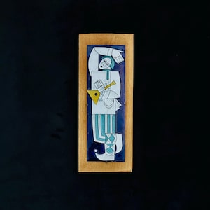 May include: A rectangular ceramic wall plaque mounted on a light wood frame. The tile features a stylised figure in white, teal, and dark blue, playing a yellow triangular stringed instrument. The figure wears a tunic, striped trousers, and boots.