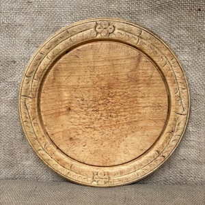 Puede incluir: Un plato redondo de madera con un acabado marrón claro y un patrón de veta visible. El borde presenta un relieve tallado con motivos de hojas y diseños de flores de cuatro pétalos situados en la parte superior e inferior del borde.