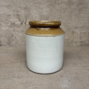 May include: A cylindrical stoneware crock featuring a smooth off-white lower body and a glossy brown glazed upper rim. A decorative band of small, raised bead-like indentations encircles the vessel at the transition between the two colours.