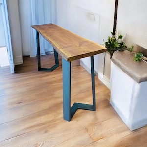 May include: A wooden console table with a natural edge top. The table has a light brown finish and is supported by teal-colored metal legs. The legs have a geometric design. The table is placed on a light-colored wooden floor.