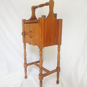 May include: A wooden side table with a small cabinet door and a handle on top. The table has four turned legs and a shelf.