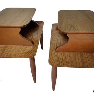 May include: Two wooden end tables with a unique design featuring a raised shelf. The tables have a light brown wood finish and tapered legs.
