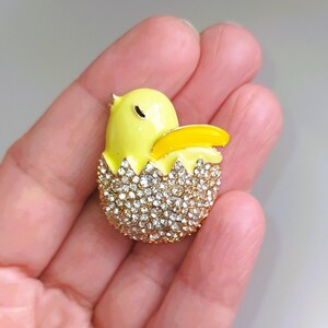 May include: A yellow chick brooch emerging from a sparkling egg. The chick is bright yellow with a yellow beak. The egg is encrusted with clear crystals, set in a gold-tone metal. A charming and whimsical accessory.