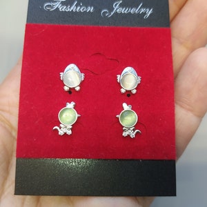 以下が含まれることがあります： 白と緑の石が付いた銀製のスタッドピアスが4組。ピアスは「Fashion Jewelry」という文字が書かれた赤いベルベットの背景に飾られています。