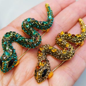 Könnte beinhalten: Zwei Broschen in Form von Schlangen, die mit Schmuck besetzt sind, eine grün und eine goldfarben mit orangefarbenen und braunen Akzenten. Die Broschen befinden sich auf einer Hand.
