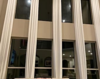 May include: A living room with tall windows draped with white curtains. A beige sofa with decorative pillows and a pink throw is in front of the windows. A wooden side table with a digital clock is on the right.