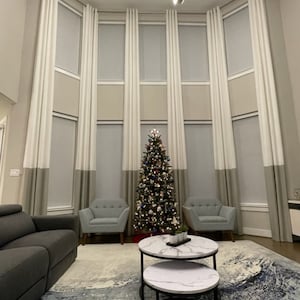 May include: A living room with a Christmas tree, two armchairs, a sofa, and two nesting coffee tables with a marble top and black metal frames. The room has large windows with white curtains and a patterned rug.