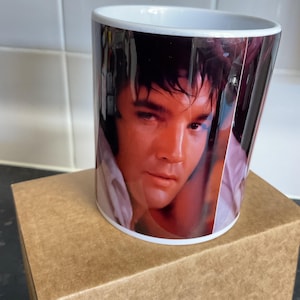 May include: White ceramic mug with a portrait of Elvis Presley. The image on the mug is a close-up of Elvis's face, with a white shirt collar visible. The mug is on a brown cardboard box.