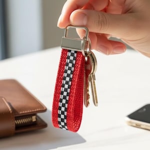 May include: Red and black checkered keychain with a silver metal clasp holding keys. The keychain is made of a woven fabric. A brown leather wallet and a smartphone are in the background.