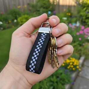 May include: A black and white checkered keychain with a silver metal loop holding several gold keys. The keychain is held in a hand against a blurred background of a garden with green grass and colorful flowers.