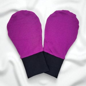 Puede incluir: Un par de mitones magenta con puños negros, sobre un fondo blanco. Los mitones están hechos de una tela suave y están diseñados para mantener las manos calientes. Los puños son de color negro contrastante.