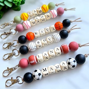 May include: A collection of personalized keychains featuring wooden letter beads spelling out names like "SAM," "MAMA," "KAYA," "ROY," "LANA," and "MAMA." Each keychain includes sports-themed beads such as baseballs, basketballs, volleyballs, footballs, and soccer balls.