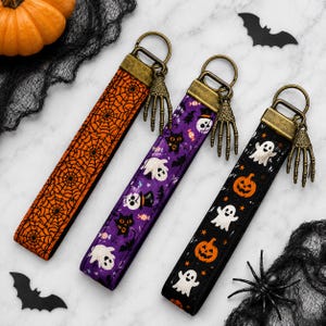 May include: Three fabric wristlet keychains with antique brass hardware and skeleton hand charms. One features an orange spiderweb pattern, one a purple background with ghosts and cats, and one a black background with ghosts and pumpkins.